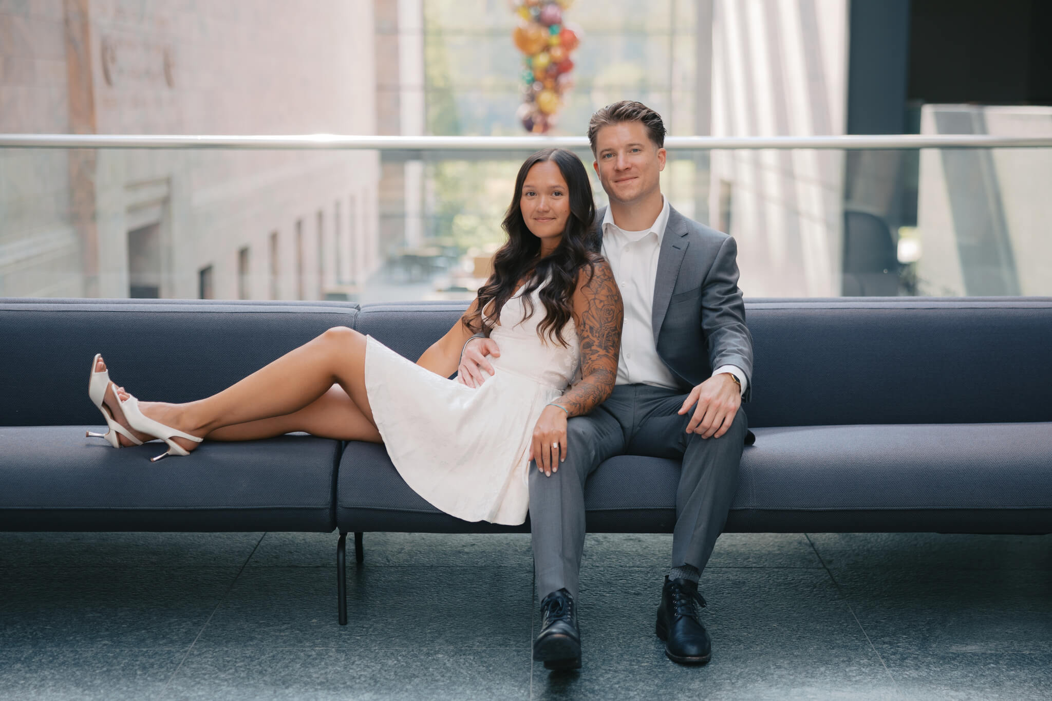 A happy couple lounge on a long bench in a lobby in a white dress and grey suit