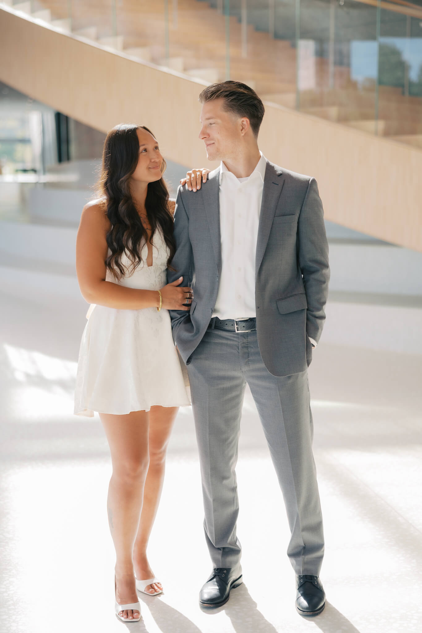 A happy woman in a white dress leans on the shoulder of her groom a they smile at each other in a large white lobby