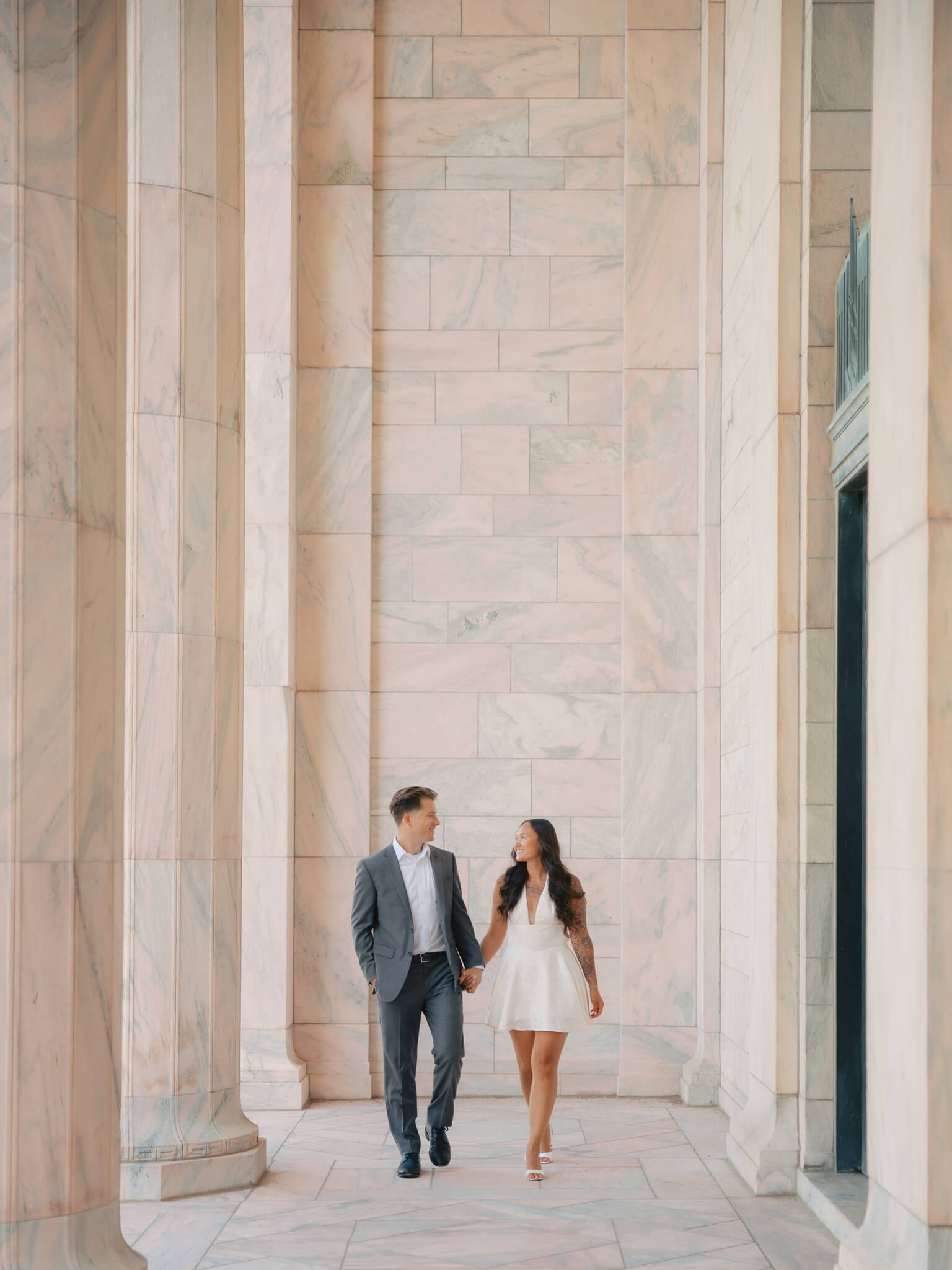 A happy couple walk holding hands through a marble colonnade at the joslyn art museum wedding venue