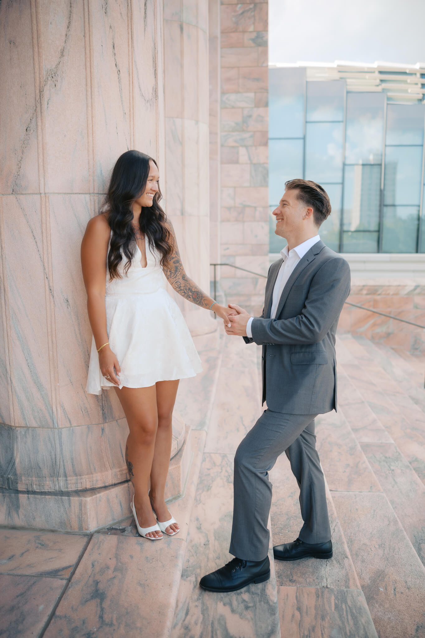 A man holds the hands of his fiancee as she leans against a marble column at the joslyn art museum wedding venue