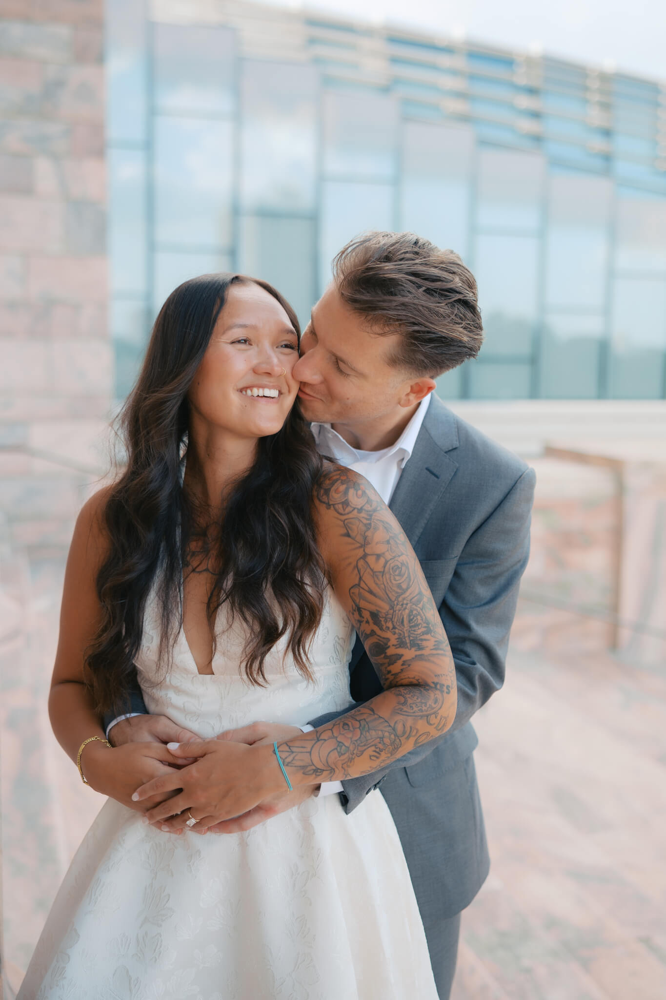 A happy woman is hugged and kissed from behind by her groom in a grey suit on the steps of the joslyn art museum wedding venue