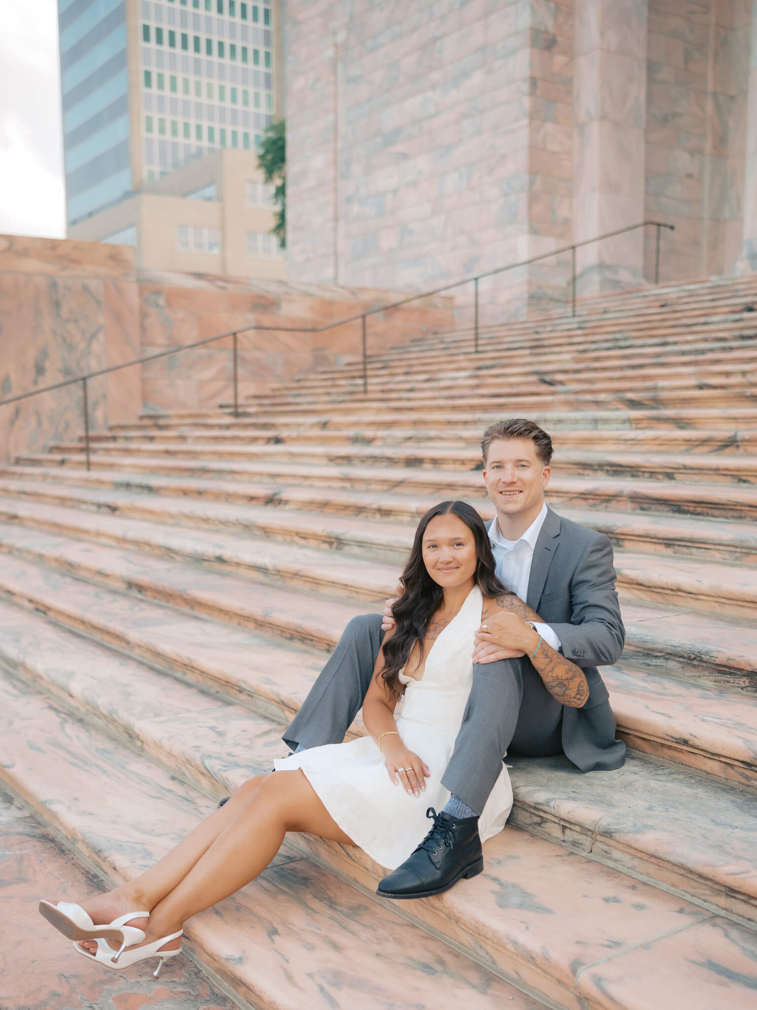A woman in white dress leans into the lap of her fiancee in a grey suit while sitting on marble steps