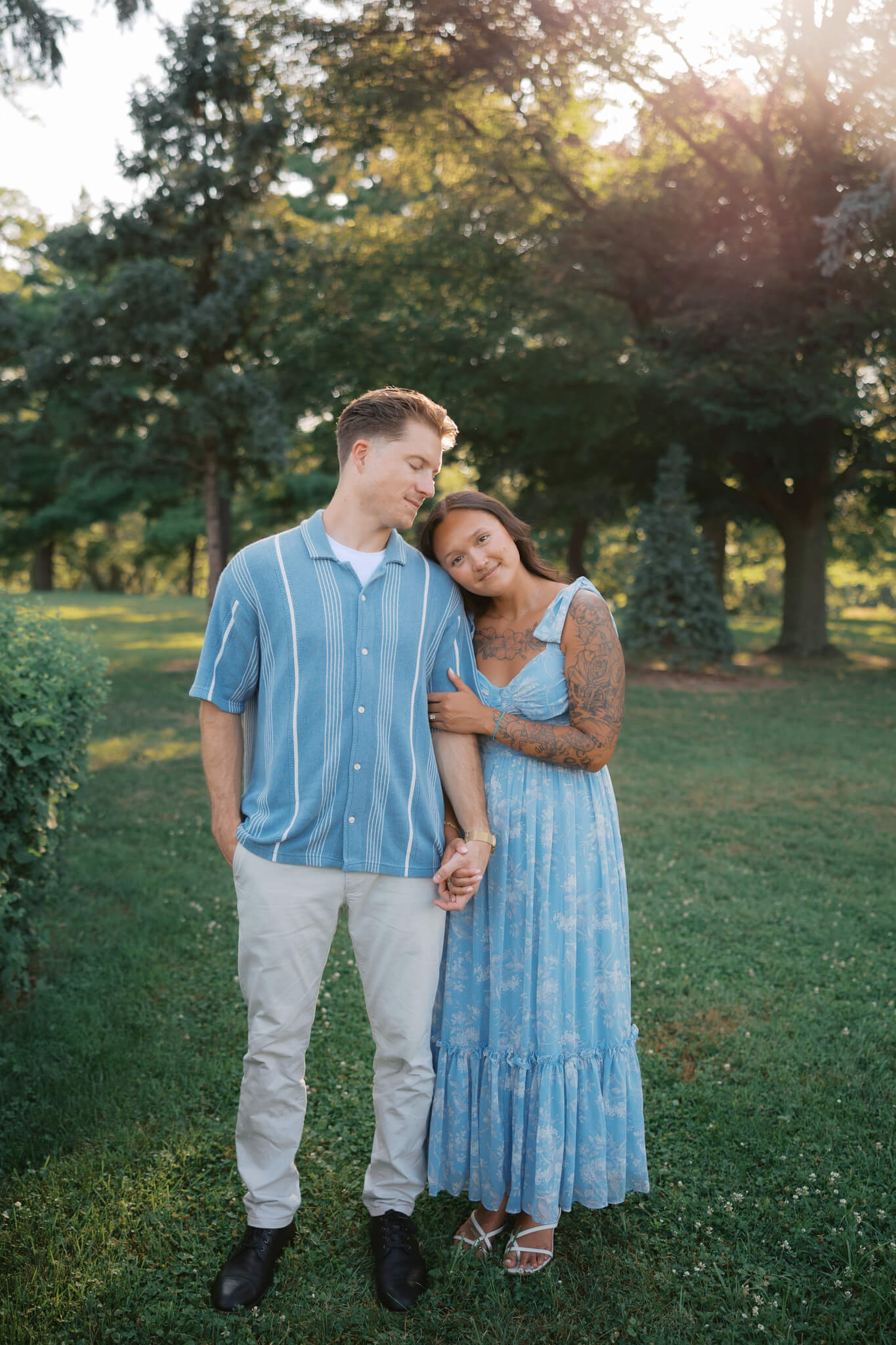 A happy engaged couple stand holding hands in blue outfits in a lawn at the joslyn art museum wedding venue